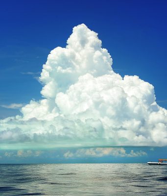 Paisaje con un gran cúmulo que se eleva hacia el cielo desde la costa, que se ve al fondo. A media distancia, un barco pequeño refuerza la majestuosidad y el tamaño enorme de la nube.