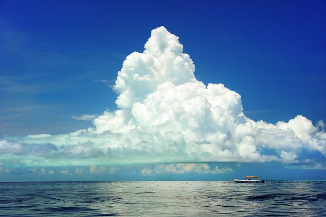 Paisaje con un gran cúmulo que se eleva hacia el cielo desde la costa, que se ve al fondo. A media distancia, un barco pequeño refuerza la majestuosidad y el tamaño enorme de la nube.