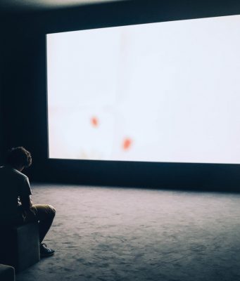 Una persona está en posición pensativa ante una gran pantalla que proyecta luz blanca.