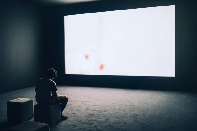 Una persona está en posición pensativa ante una gran pantalla que proyecta luz blanca.