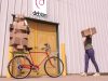 Entrada de una factoría que tiene un cartel de Debian sobre la puerta de entrada. Una mujer trae un paquete. Parece haber aparcado una bici con más paquetes, sujetos con un pulpo (elástico).