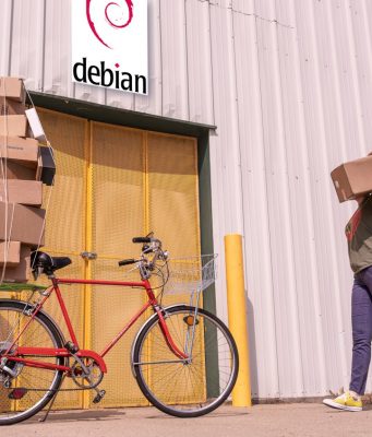 Entrada de una factoría que tiene un cartel de Debian sobre la puerta de entrada. Una mujer trae un paquete. Parece haber aparcado una bici con más paquetes, sujetos con un pulpo (elástico).